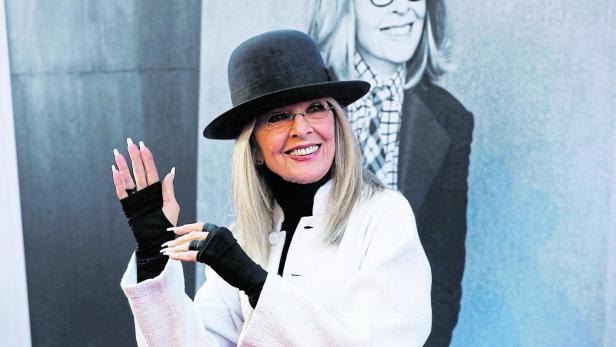 Diane Keaton lächelt mit Hut und weißen Mantel vor einem großen Porträt.