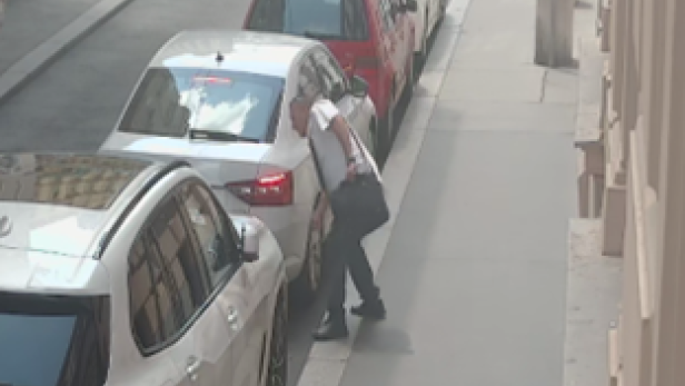 Ein Mann in weißem T-Shirt steht auf einem Gehsteig und scheint einen Reifen der parkenden Autos aufzustechen.