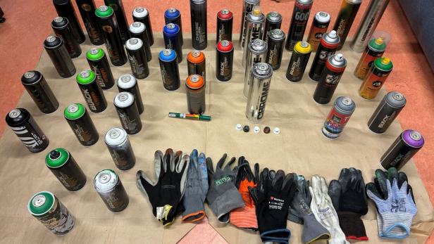 Mehrere Farbsprühflaschen sind auf einem Tuch neben unterschiedlichen Handschuhpaaren aufgestellt.