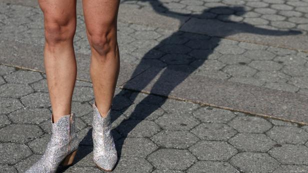 Glitzernde Stiefeletten an nackten Beinen auf Pflastersteinen, daneben ein großer Schatten mit ausgebreiteten Armen.