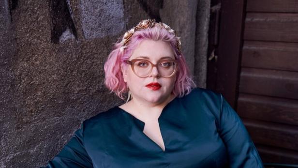 Bobby Herrmann-Thurner sitzt auf einer Bank, trägt einen dunkelblauen Jumpsuit und rosa Haare.