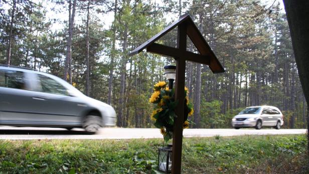 46-17423326 Ein Holzkreuz steht am Straßenrand und zwei Autos fahren im Hintergrund vorbei.
