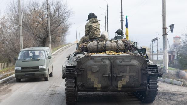Kupiansk Ein Soldat in Tarnuniform sitzt auf einem Panzer, der eine schmale Straße entlangfährt, daneben ein grüner Transporter.