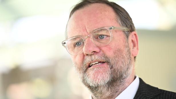 Ein älterer Mann mit Brille, grauem Bart und Anzug spricht mit ernstem Gesichtsausdruck.