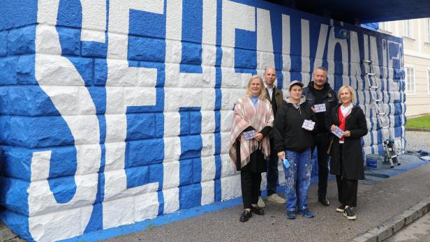 Eine Gruppe an Menschen steht unter einer Brücke, die Wände wurden frisch mit blauer Farbe und weißer Schrift versehen.