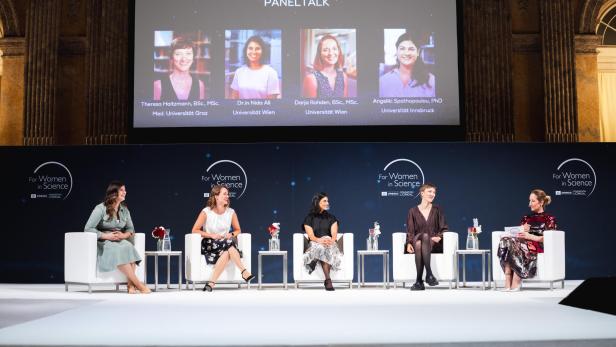 Fünf Frauen sitzen auf einer Bühne bei einer Podiumsdiskussion, im Hintergrund eine Leinwand mit Fotos und Namen der Teilnehmerinnen.