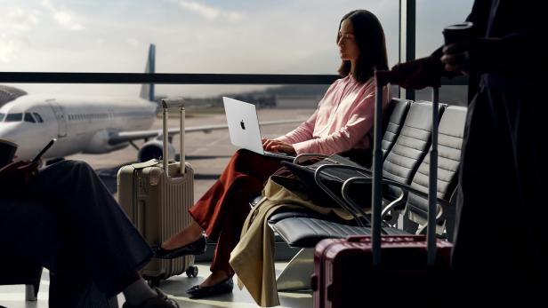 Frau arbeitet mit Laptop am Flughafen, Koffer neben ihr, Flugzeug im Hintergrund.