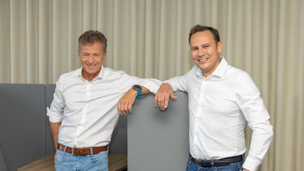 46-219062066 Zwei Männer in weißen Hemden und Jeans stehen lächelnd nebeneinander an einer grauen Trennwand.