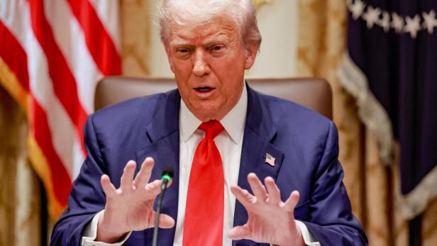 Donald Trump gestikuliert vor US- und Sternenbannerflaggen mit roter Krawatte und blauem Anzug.