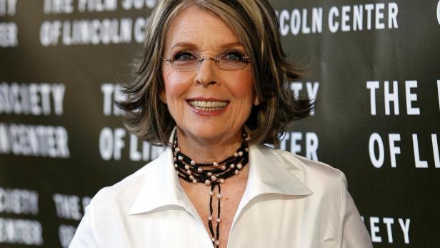 Diane Keaton lächelt in weißer Bluse, schwarzem Gürtel und Perlenkette vor einem schwarzen Hintergrund.