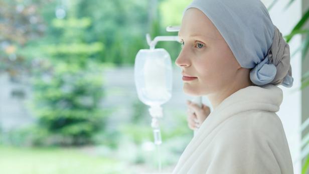 Eine Frau mit Kopftuch und Bademantel steht mit Infusion am Fenster und blickt nach draußen.