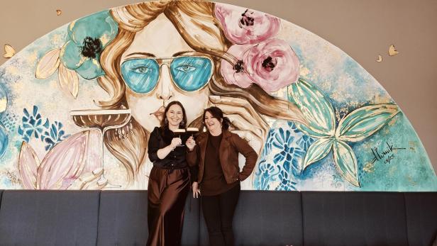Zwei Frauen stehen lachend vor einer bunten Wandmalerei mit Blumen und einer Frau mit blauer Sonnenbrille.