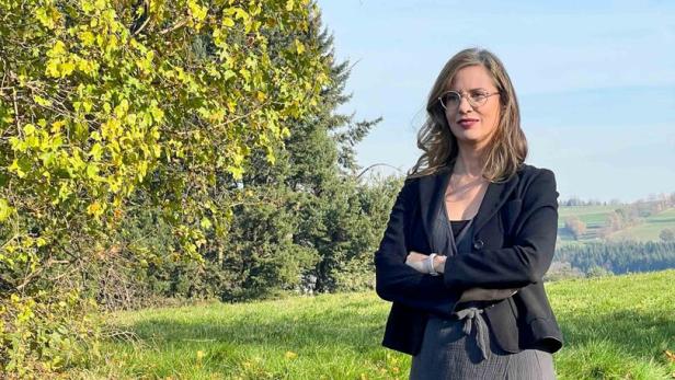 46-219479256 Eine Frau mit Brille, in Kleid und Blazer, steht auf einer Wiese mit Bäumen im Hintergrund.