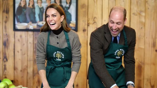 Kate und William Kate und William lachen beim Teig ausrollen, beide tragen grüne Schürzen mit dem Aufdruck "Long Meadow Farm".