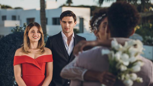 Aus dem Film Culpa Nuestra: Zwei elegant gekleidete Personen stehen lächelnd nebeneinander, während im Vordergrund ein Paar sich umarmt und Blumen hält.