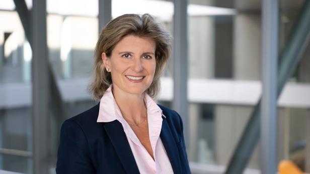 Eine lächelnde Frau mit Blazer und Bluse steht vor einem modernen Gebäude.