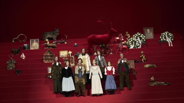 Acht Personen in Tracht stehen auf roten Stufen, umgeben von Jagdtrophäen, Blumenkränzen und Dekorationsgegenständen.