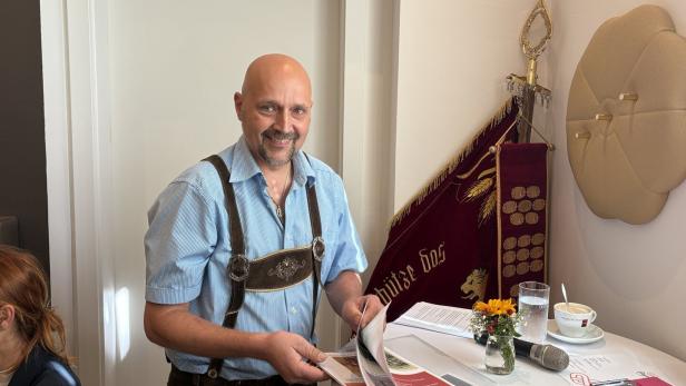 46-219479243 Ein Mann in Tracht steht lächelnd an einem Tisch mit Unterlagen und einer Fahne.