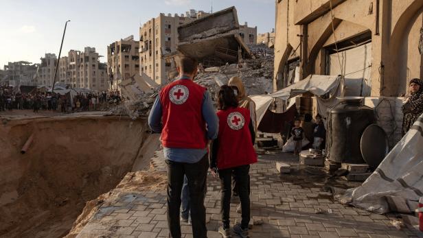 Red Cross inspects search operation for Israeli hostages bodies Zwei Helfer des Roten Kreuzes stehen vor zerstörten Gebäuden und einem großen Krater, während Menschen im Hintergrund zusehen.