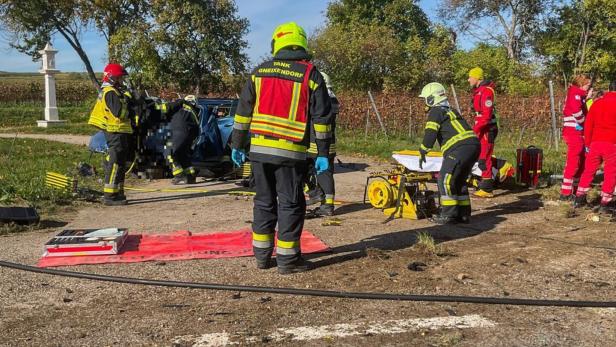 Mehrere Feuerwehr- und Rettungskräfte arbeiten gemeinsam an einem Unfallauto auf einer Landstraße.
