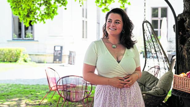Talk mit Schauspielerin Ronja Forcher Eine Frau mit dunklen Haaren steht lächelnd im Schatten eines Baumes neben einem Hängesessel und einem Korb mit Obst.