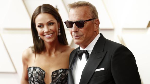 Kevin Costner und Christina Baumgartner Kevin Costner posiert mit Christina Baumgartner in einem trägerlosen Kleid und trägt eine Sonnenbrille und einen Smoking.