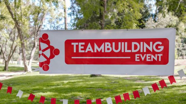 Fünf weiße Bowlingkegel mit roten Ringen auf Gras, darüber ein Banner mit rotem Schriftzug 'TEAMBUILDING EVENT' und roter Grafik, im Hintergrund Bäume.