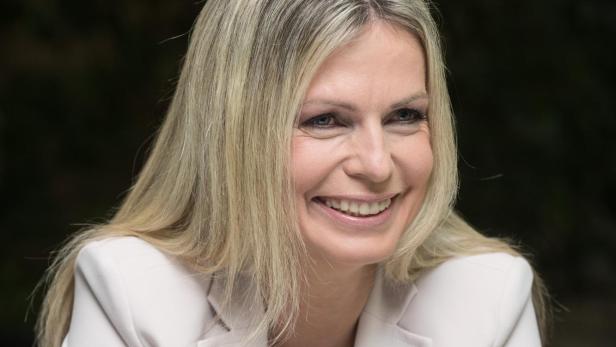 Claudia Reiterer Eine lächelnde Frau mit langen blonden Haaren trägt einen hellen Hosenanzug und sitzt mit gefalteten Händen.