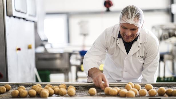 Weinbergmaier Produktion Ein Mann in weißem Kittel und Haarnetz sortiert Kartoffeln auf einem Förderband in einer Produktionshalle.