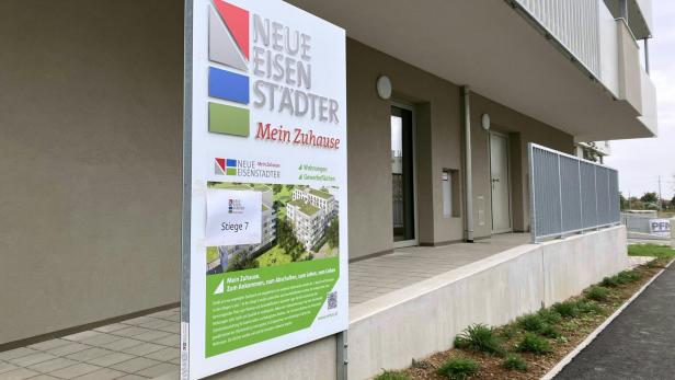 Ein Schild mit dem Schriftzug „Neue Eisenstädter Mein Zuhause“ steht vor einem modernen Wohngebäude mit Eingängen und bepflanztem Rand.