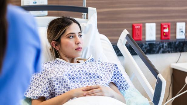 Ein Verlust des noch ungeborenen Kindes in der frühen Schwangerschaft führt bei Frauen häufig zu Schuld- und Versagensgefühlen. Eine junge Frau liegt mit gefalteten Händen in einem Krankenhausbett und blickt nachdenklich zur Seite.