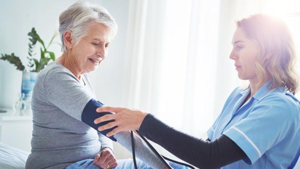 Ältere Frau sitzt, während eine Pflegekraft in blauer Arbeitskleidung ihren Blutdruck misst. Ältere Frau sitzt, während eine Pflegekraft in blauer Arbeitskleidung ihren Blutdruck misst.