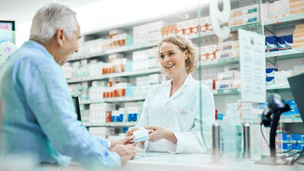 Apothekerin in weißem Kittel übergibt einem Kunden ein Medikament in einer Apotheke mit Regalen voller Arzneimittel im Hintergrund. Apothekerin in weißem Kittel übergibt einem Kunden ein Medikament in einer Apotheke mit Regalen voller Arzneimittel im Hintergrund.