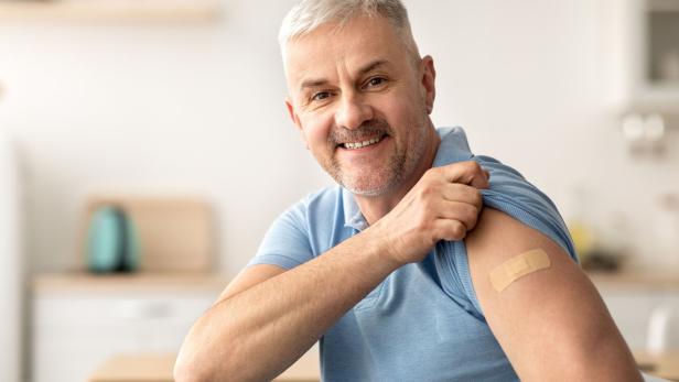 Mann in blauem Poloshirt zeigt Pflaster auf Oberarm, sitzt in heller Küche mit unscharfem Hintergrund.