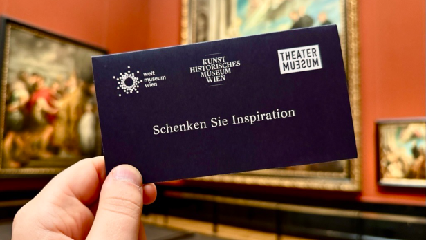 Hand hält dunkelblauen Gutschein mit den Logos von Weltmuseum Wien, Kunsthistorisches Museum Wien und Theatermuseum sowie dem Text 'Schenken Sie Inspiration' vor Gemälden in Museumssaal.