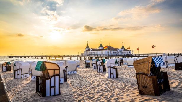 Strand mit mehreren Strandkörben in verschiedenen Farben und einer Seebrücke mit Gebäude im Hintergrund bei Sonnenuntergang.