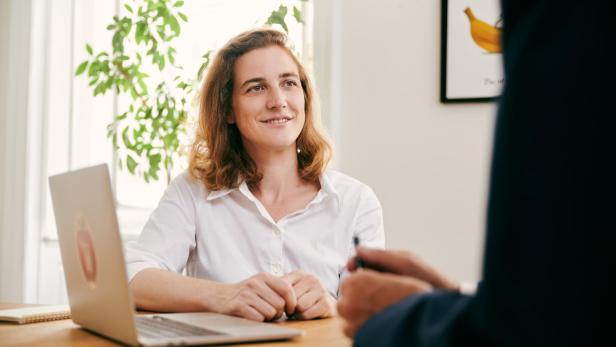 Frau mit weißem Hemd sitzt an einem Holztisch vor einem Laptop, im Hintergrund eine grüne Pflanze und ein Bild mit einer Banane an der Wand.