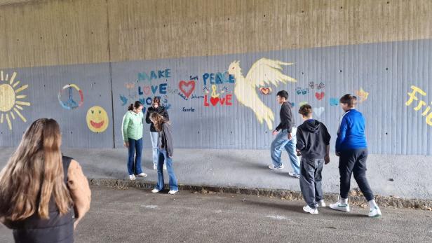 Mehrere Jugendliche stehen vor einer bemalten Wand mit Friedenssymbolen, einer Taube, einer Sonne und positiven Botschaften.