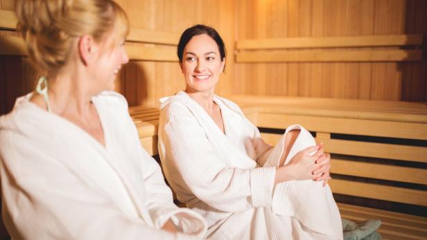 Zwei Frauen im Bademantel sitzen entspannt in einer Sauna mit Holzverkleidung und unterhalten sich.