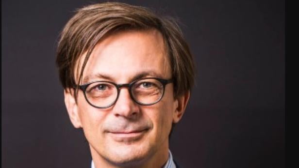 Ein Mann mit Brille, Anzug und Krawatte vor einem dunklen Hintergrund.