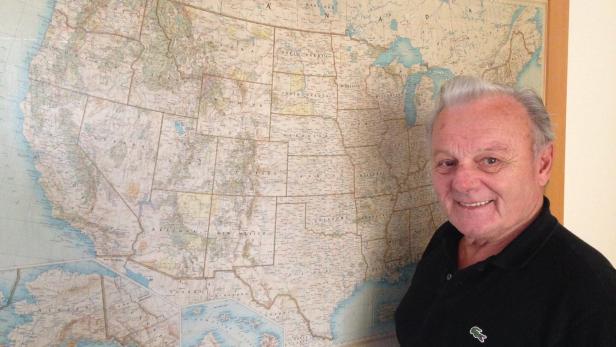 Ein älterer Mann mit grauen Haaren steht lächelnd vor einer großen Landkarte der USA an der Wand.