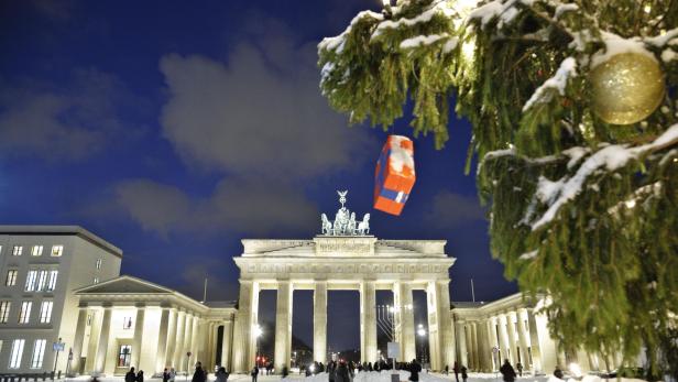 Brandenburger Tor bei Nacht mit Schnee, davor Menschen und ein Weihnachtsbaum mit goldener Kugel im Vordergrund.