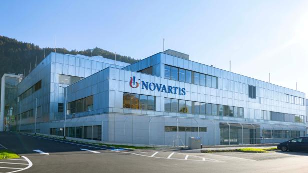 Modernes Bürogebäude mit Glasfassade und dem Schriftzug 'Novartis' an der Frontseite, umgeben von Parkplätzen und Bergen im Hintergrund.