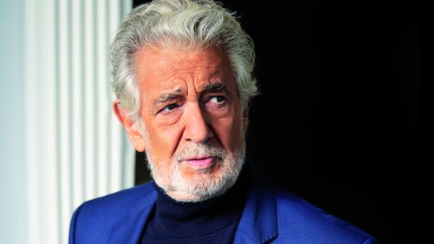 Älterer Mann mit grauen Haaren und Bart trägt einen dunkelblauen Anzug und schwarzen Rollkragenpullover vor schwarzem Hintergrund.