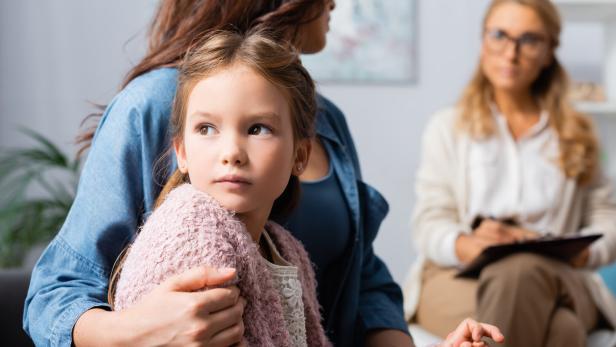 Ein Mädchen sitzt auf dem Schoß einer Frau und wird von ihr umarmt, während im Hintergrund eine weitere Frau Notizen macht.