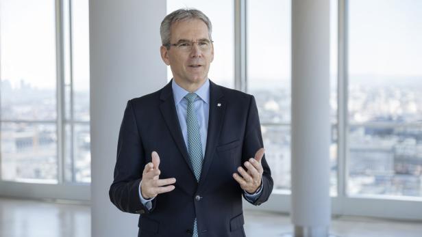 Mann in dunklem Anzug mit hellblauem Hemd und gemusterter Krawatte steht vor großen Fenstern mit Stadtblick, gestikuliert mit beiden Händen.