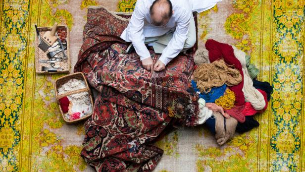Vogelperspektive: Ein Mann hockt am Boden, vor ihm ein Teppich, neben ihm Wolle und Flickzeug. Er arbeitet am Perser mit klassischem Muster.