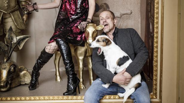 46-219701361 Eine Person mit roter Perücke und auffälligem Outfit steht neben einem Mann mit Hund auf dem Schoß in einer dekorativen Kulisse.