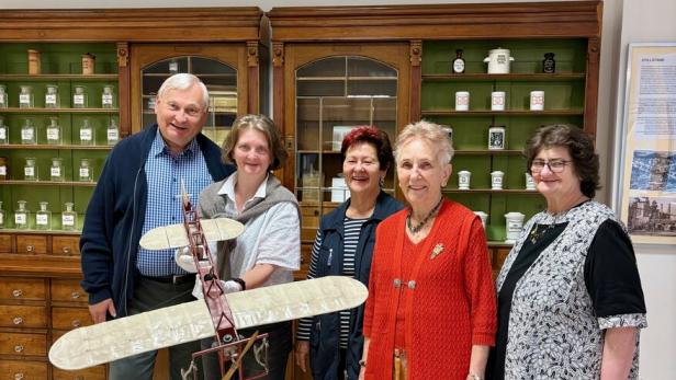 Fünf Personen stehen in einem Raum mit alten Apothekerschränken, eine Person hält ein Modellflugzeug.