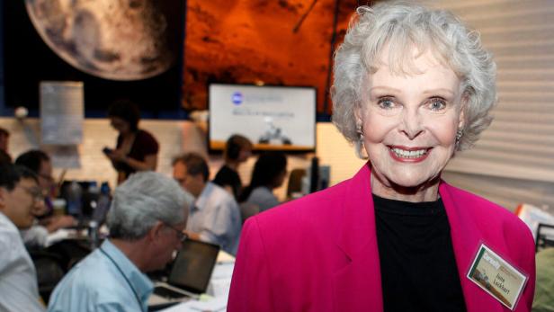 FILE PHOTO: Actress Lockhart, best know for her TV roles in "Lost in Space" and "Lassie", poses for a photograph in the newsroom at NASA's Jet Propulsion Lab in Pasadena June Lockhart lächelt vor einem Hintergrund mit Planetenbildern und einem Raum voller Menschen an Computern.